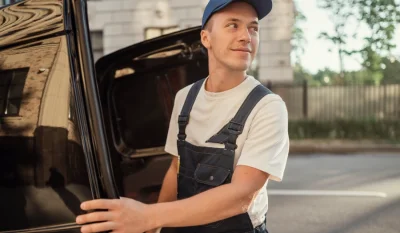 Mecánico con gorra azul y mono abriendo la puerta de un coche oscuro a pleno sol