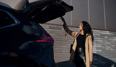 Mujer con abrigo beige abriendo el maletero de un coche oscuro junto a un muro de ladrillo