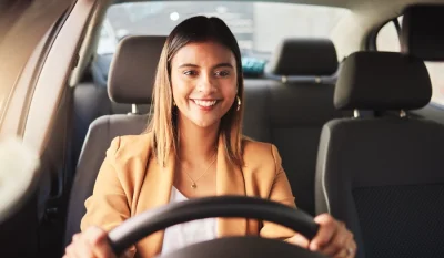 Mujer sonriente con chaqueta beige al volante de un coche, con las manos en el volante