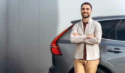 Hombre con camisa beige de brazos cruzados junto a un coche gris con luz trasera roja