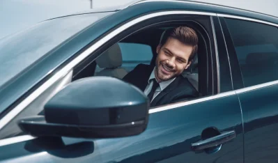 Hombre de traje sonriendo al volante de un coche azul oscuro, visto por la ventanilla