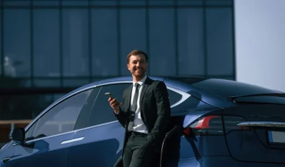 Hombre de traje con el móvil en la mano junto a un coche azul marino frente a un edificio de cristal