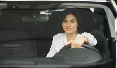 Mujer con camisa blanca al volante de un coche oscuro, mirando de frente