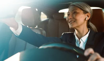 Mujer con auriculares y chaqueta oscura conduciendo y sonriendo, con la mano en el volante