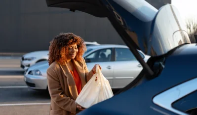 Mujer con bolsa blanca junto al portón abierto de un coche azul marino, con más coches al fondo