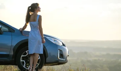 Mujer con vestido azul junto a un coche azul, con un paisaje de campo al fondo