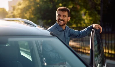Hombre sonriente apoyado en la puerta de un coche gris oscuro junto a una barandilla