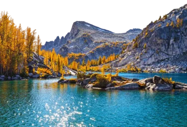 Lago turquesa entre lariches dorados y montañas rocosas, con el sol reflejado en el agua
