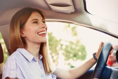 Mujer sonriente conduciendo con una mano en el volante y la luz del sol entrando por el parabrisas