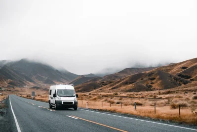 Furgoneta blanca circulando por una carretera recta entre colinas áridas con niebla