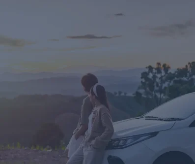 Mujer junto a un coche blanco mirando un paisaje de bosque y colinas al atardecer