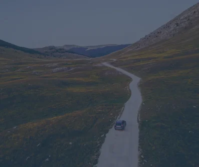 Vista aérea de una carretera sinuosa entre prados verdes y montañas, con un coche