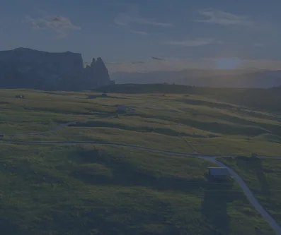 Paisaje de prados verdes y picos rocosos bajo un cielo nublado, con un camino al fondo