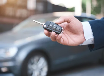 Mano con camisa blanca mostrando una llave de coche frente a un coche gris desenfocado