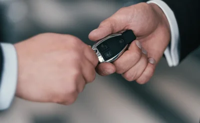Mano de traje entregando un mando de llave de coche a otra mano sobre fondo gris difuminado