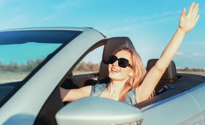 Mujer sonriente con el brazo en alto desde un descapotable claro y gafas de sol