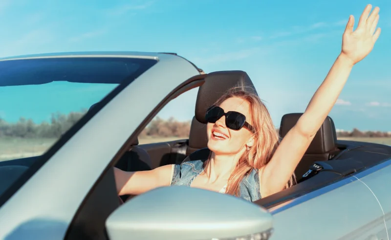 Mujer sonriente con el brazo en alto desde un descapotable claro y gafas de sol